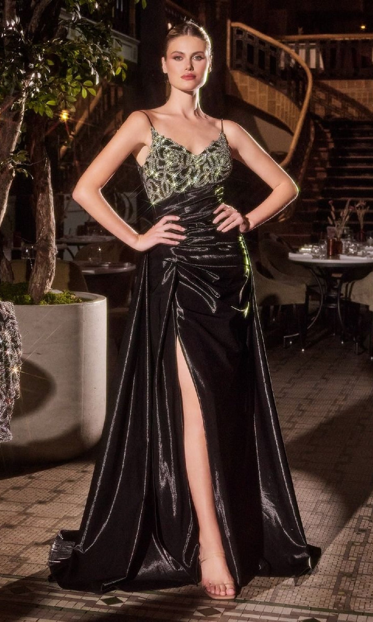 Woman poses outdoors in the Ladivine J882 Jacket Long Black Formal Dress, featuring a beaded bodice and thigh-high slit, exuding confidence with hands on hips.